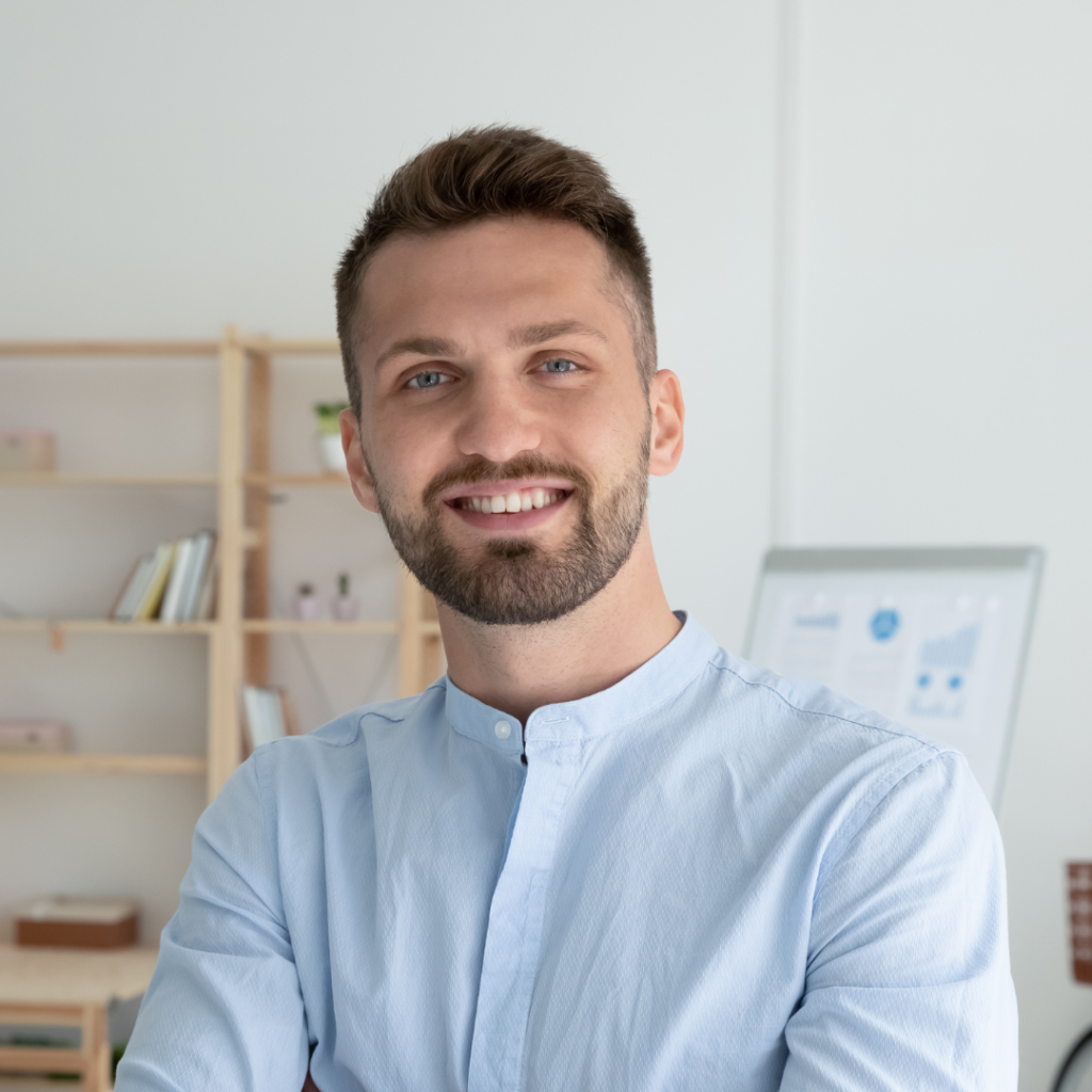 Smiling young man in a light blue shirt, standing in a modern workspace.